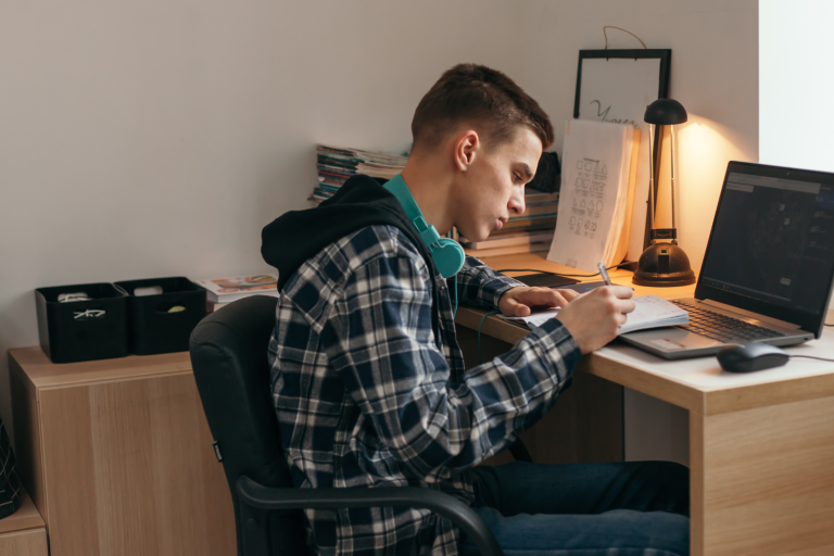 Teenage boy writing on notepad at desk in front of laptop