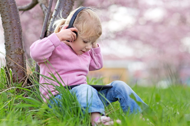 Toddler girl sitting by a tree looking at device with headphones on